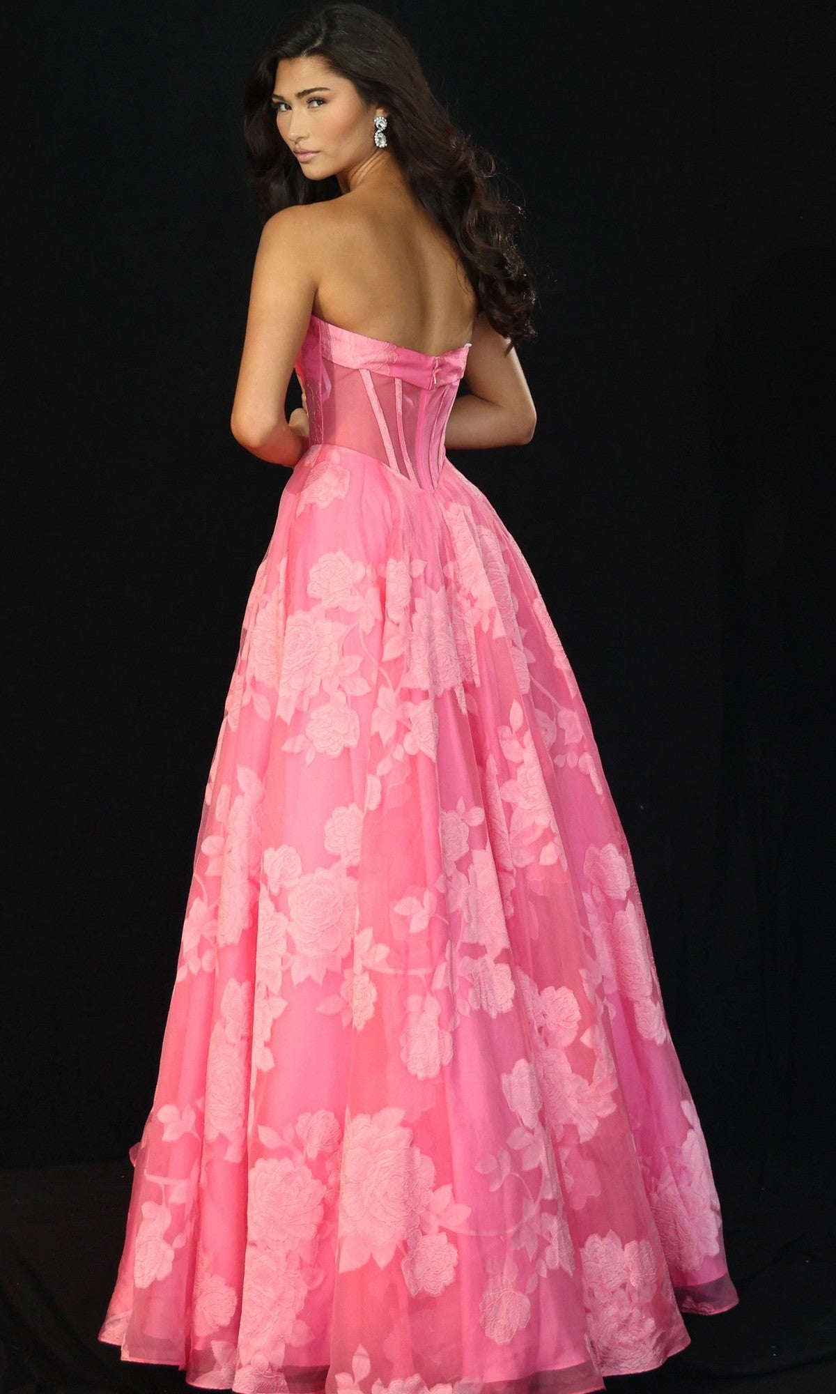 A woman wearing the Long Prom Dress: Jovani 47327, featuring a strapless floral bodice and floor-length pink skirt, stands against a black background, looking over her shoulder.